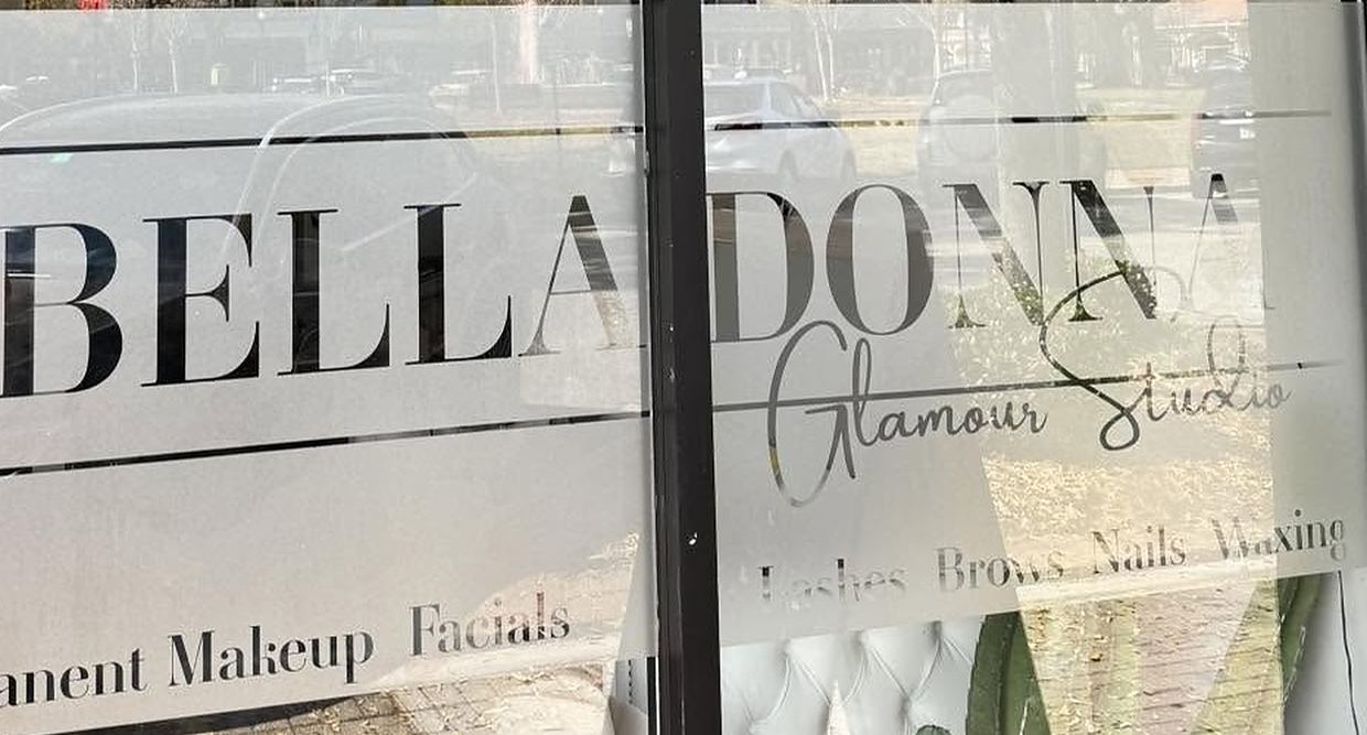 Belladonna Glamour Studio storefront window in downtown Winter Haven, with the studio name and service list etched into the glass: permanent makeup, facials, lashes, brows, nails, waxing. Street traffic reflects in the window and white tufted seating is visible inside.